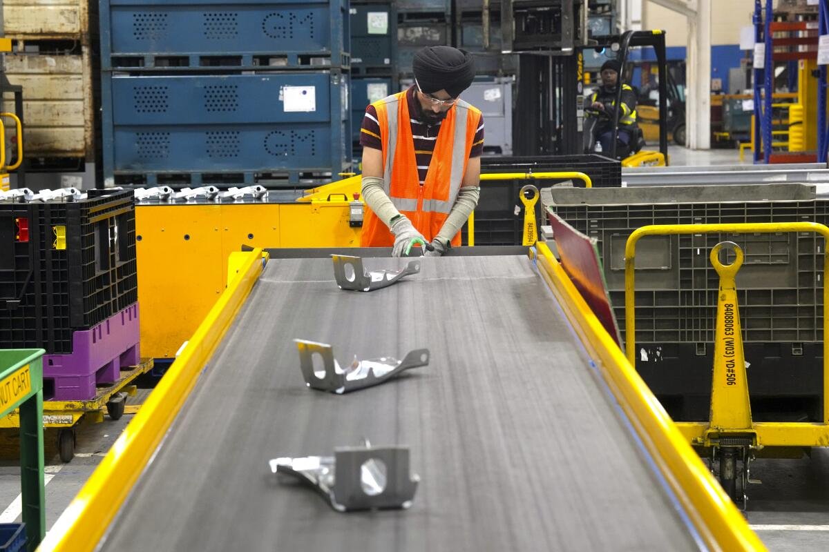 A worker in an orange vest stands at the end of a conveyor belt with metal parts on it