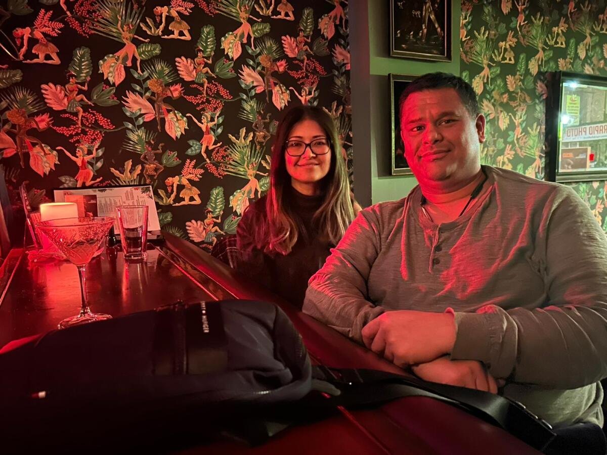 A woman with brown hair and glasses and a man are seated at a counter in a bar