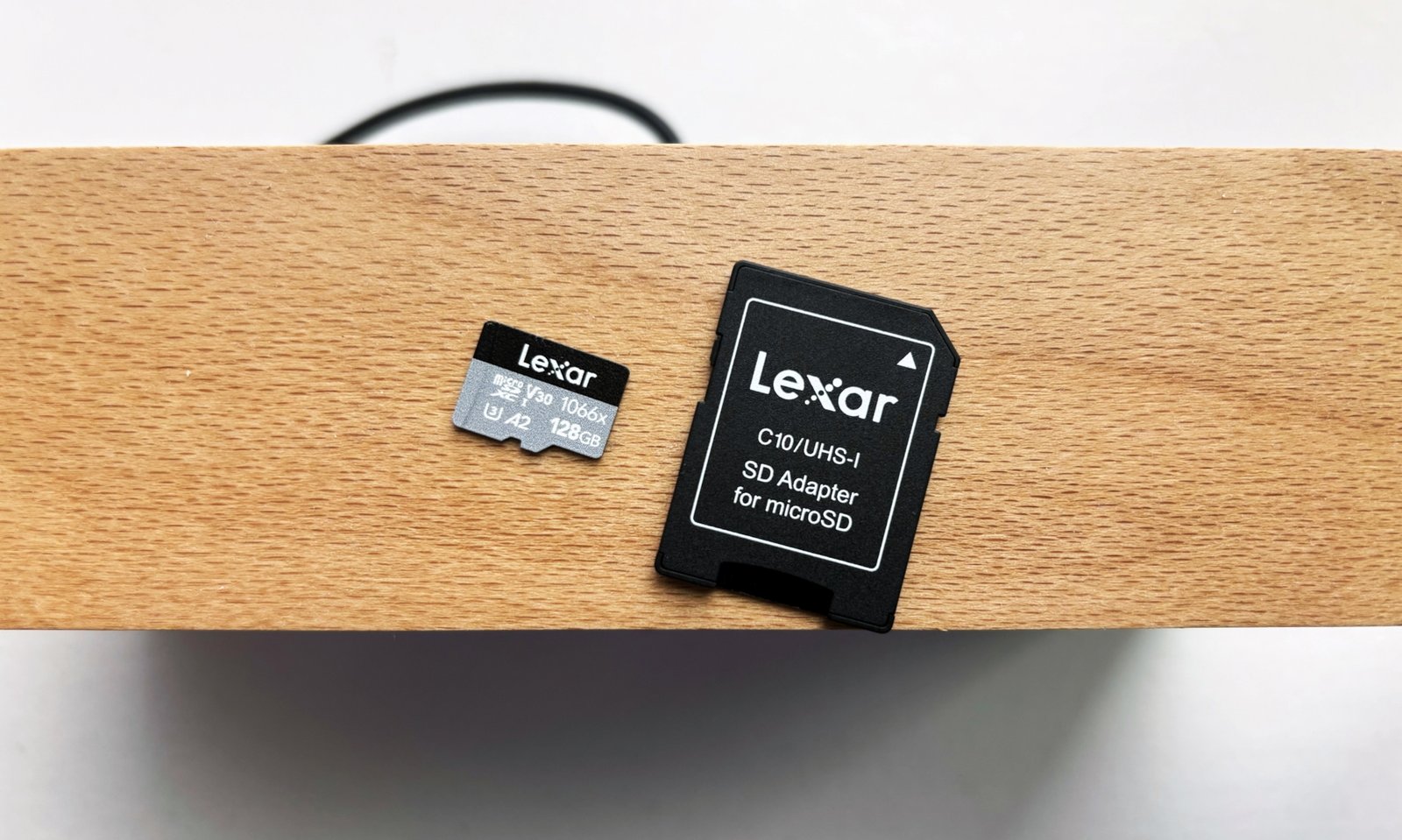 A silver and black Lexar Professional 1066x microSD card and its black SD card adapter rest on top of a brown wooden shelf above a white window sill.